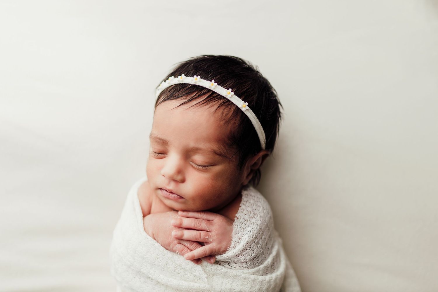Peaceful newborn wrapped in white swaddle with delicate headband against light backdrop.