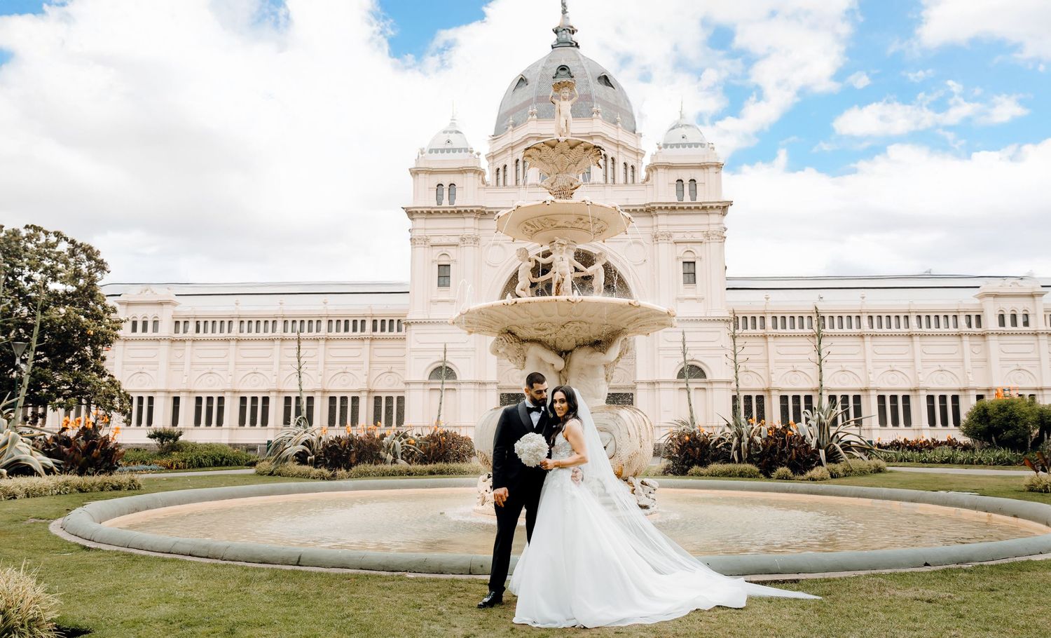 A couple in wedding attire poses in front of the Royal Exhibition Building in Melbourne with its grand dome and fountain.