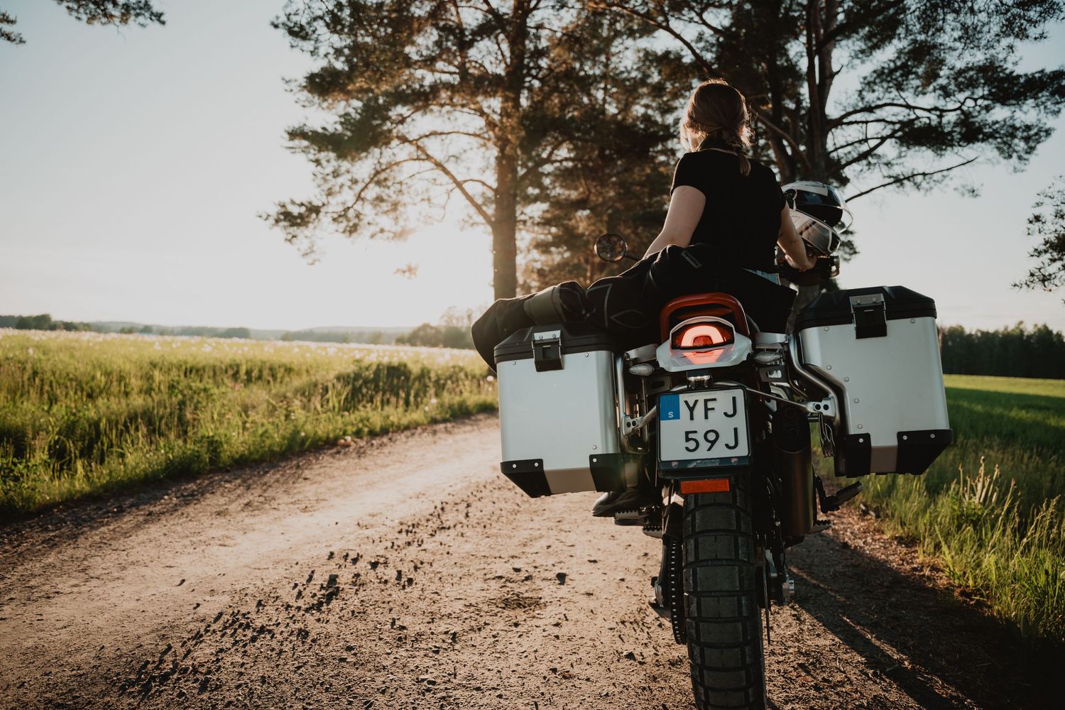 Kvinna på motorcykel blickar ut över grusväg och fält i solnedgång. Fotograf Gabriel Alenius, Falun