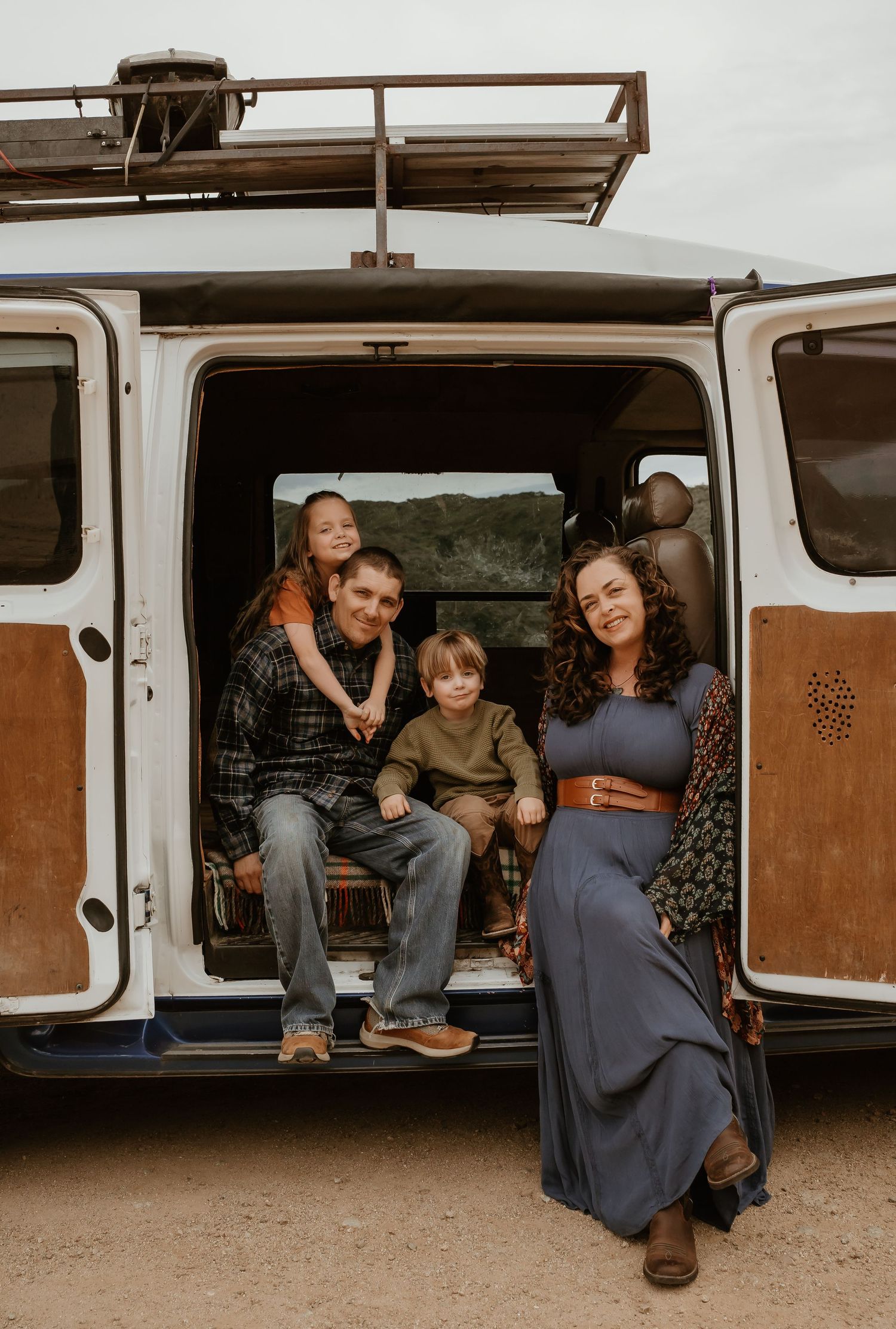 A vintage two-tone van with a roof rack parked on a dirt path, with occupants seated in the open back door.