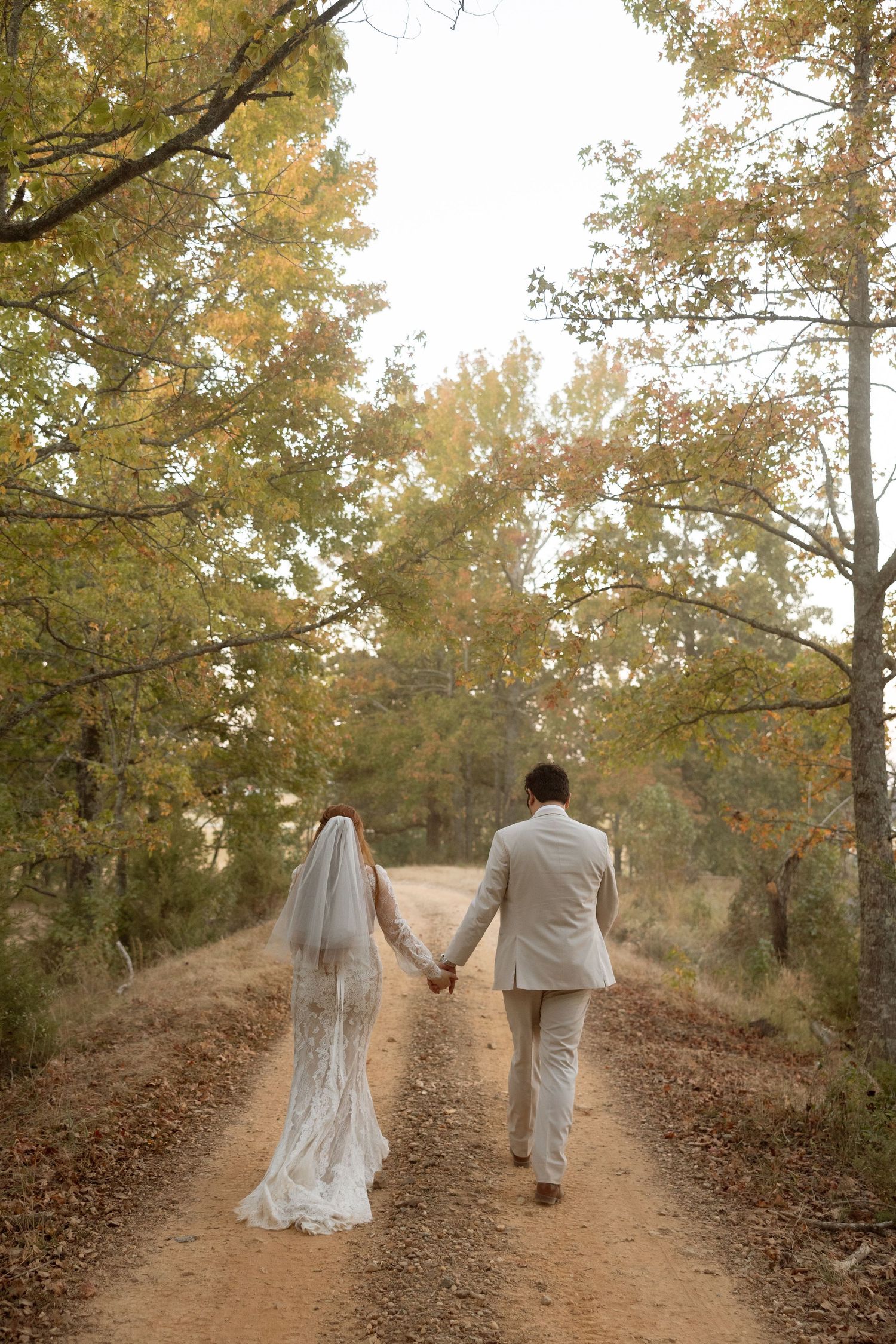 A couple in wedding attire walks down a tree-lined dirt path surrounded by autumn foliage.