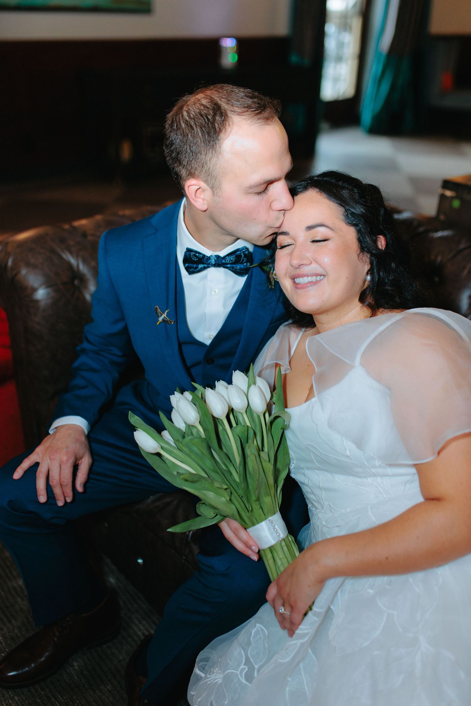 A tender wedding moment with couple seated on leather couch as bride holds white tulip bouquet at intimate indoor venue.
