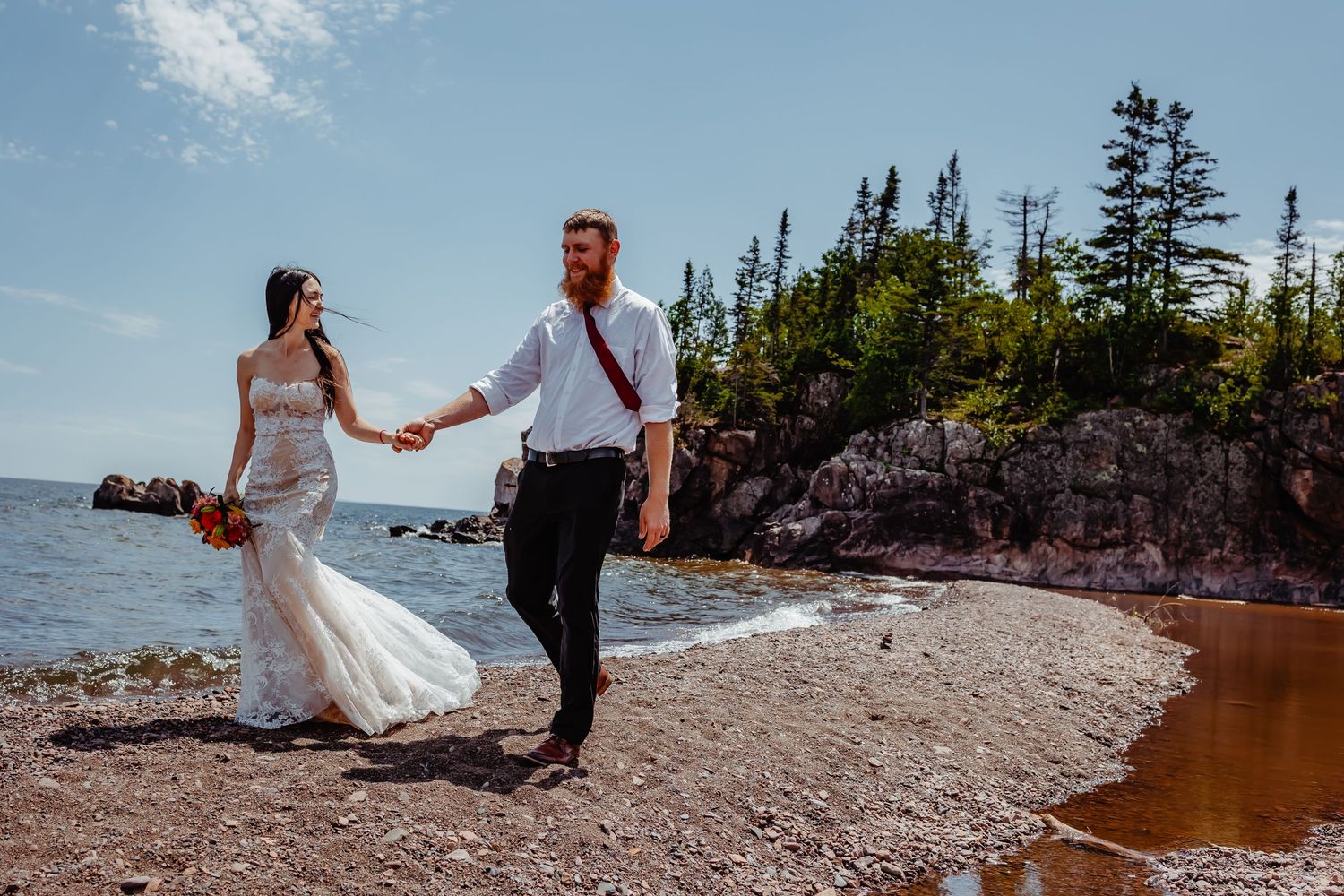 A couple walking hand in hand on a rocky beach with evergreen trees in the background.