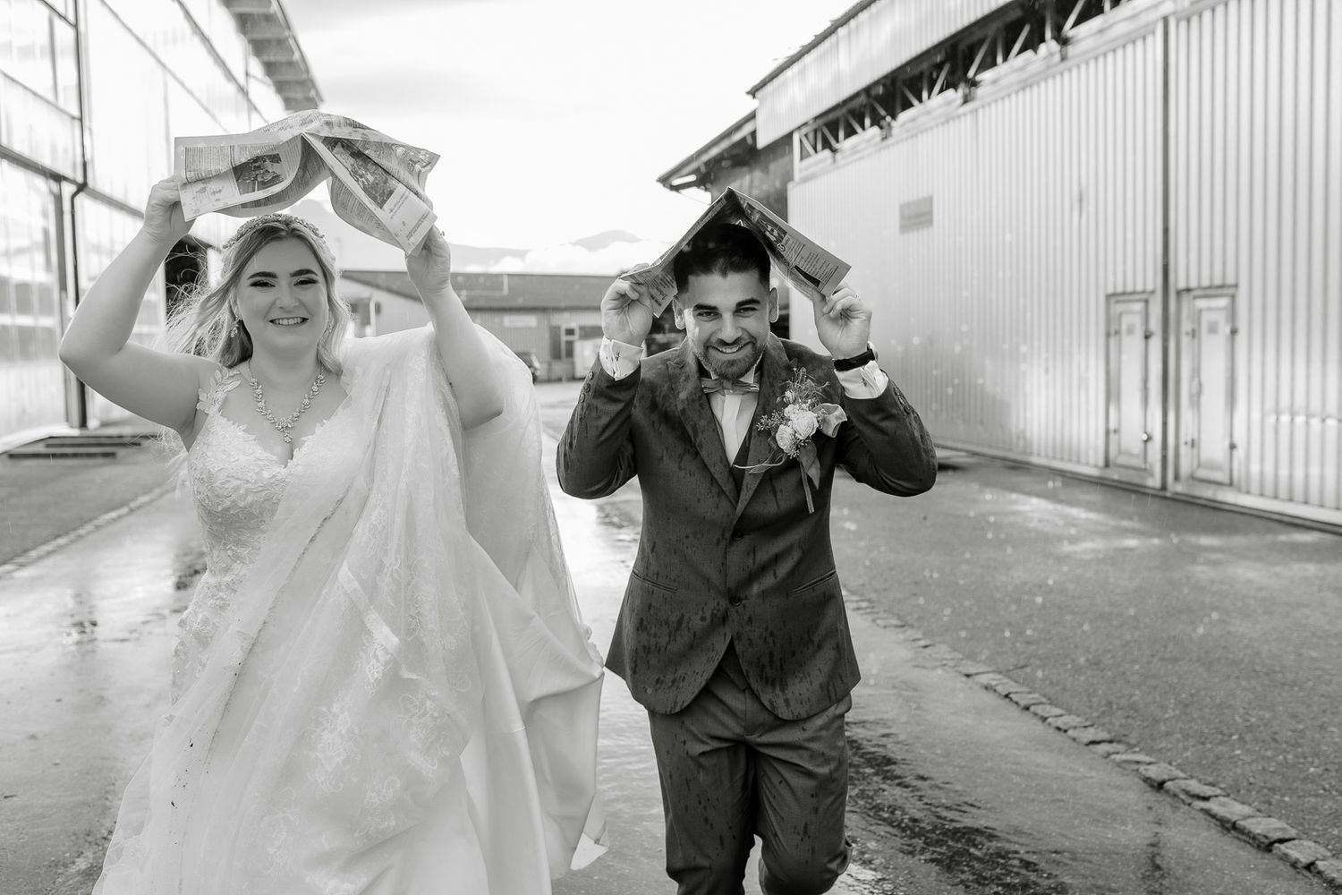 Black and white photo of a bride and groom laughing while holding objects over their heads in the rain.