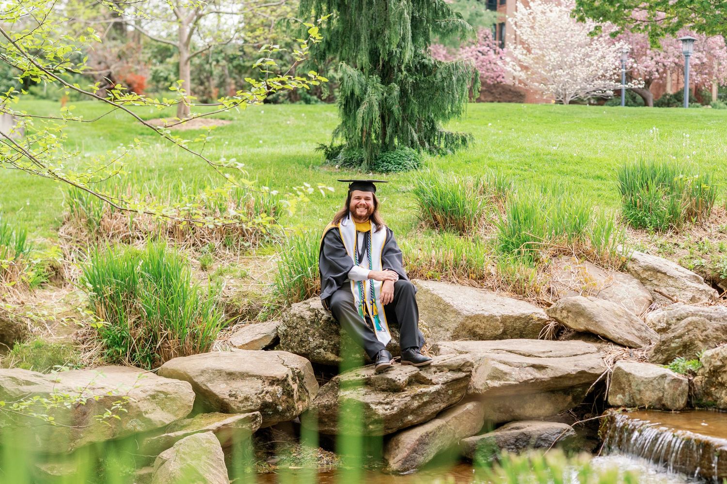 A graduate poses on rocks near a peaceful garden pond wearing academic regalia.