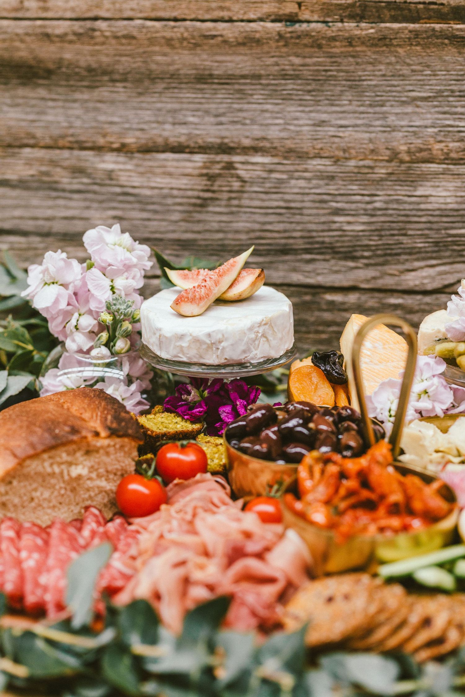 Colorful charcuterie board with meats, cheeses, fruits and flowers on rustic wooden surface.
