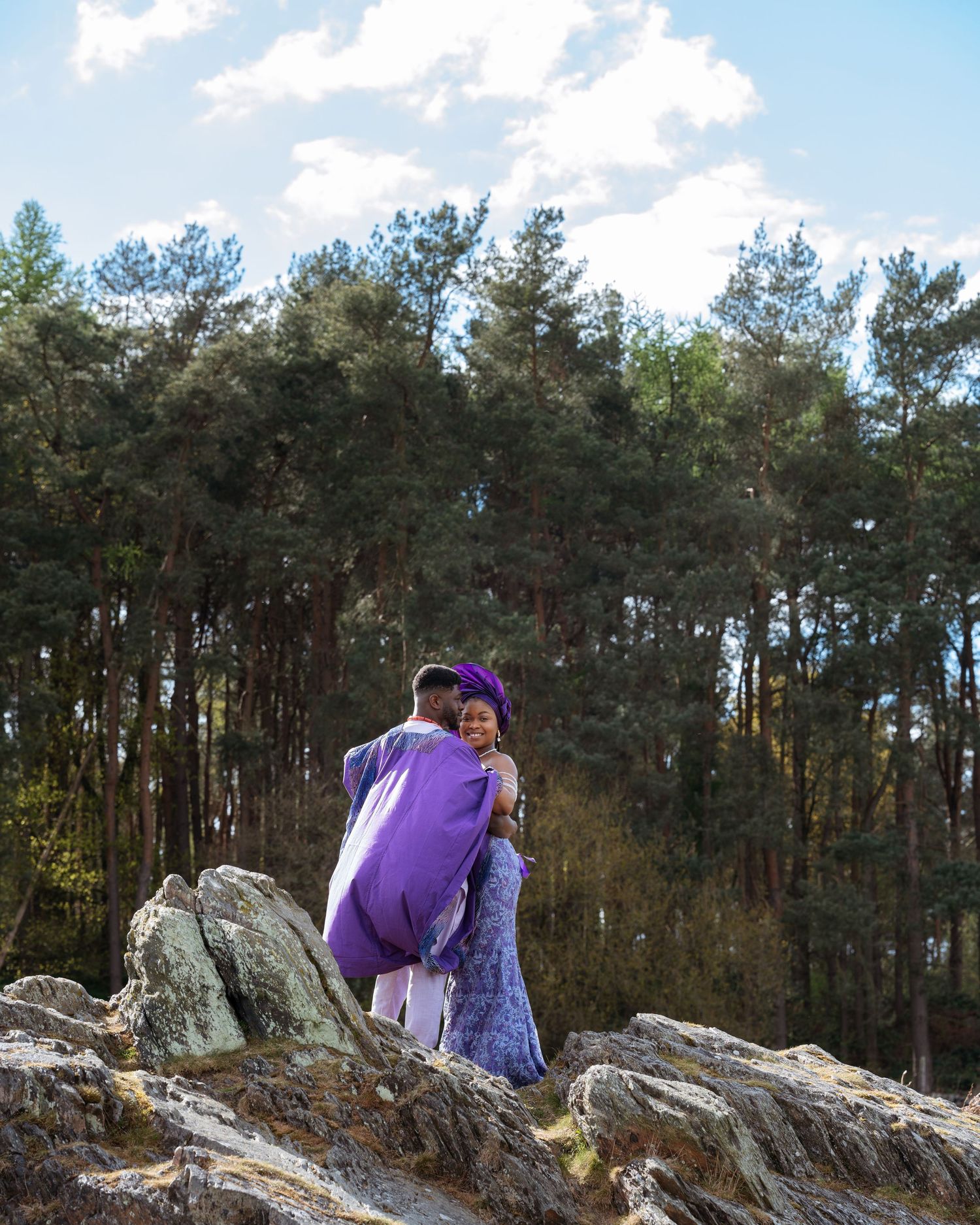 Person wearing purple and grey outdoor apparel sits atop rocky outcrop with pine forest in background.