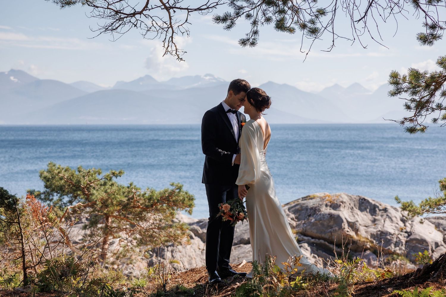 Newlyweds share an intimate moment on rocky coastal cliffs with mountains in the background.