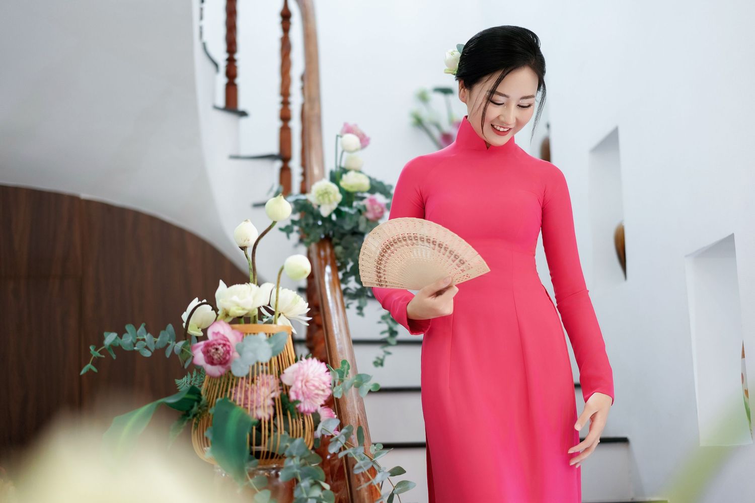 Traditional red ao dai dress worn against floral decorations and wooden staircase.