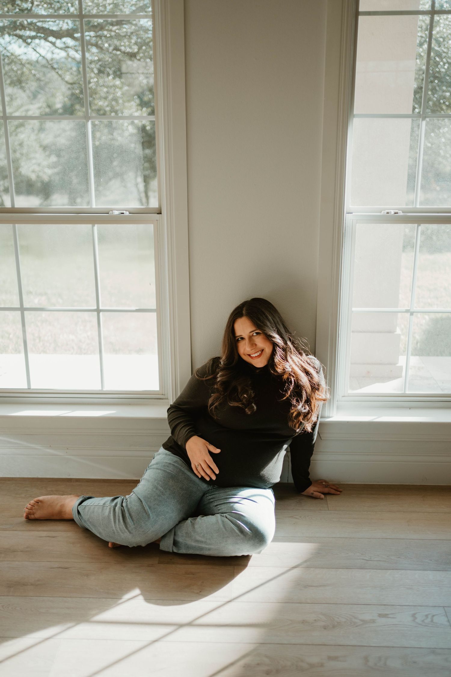 Natural light portrait of woman sitting on hardwood floor between two bright windows.
