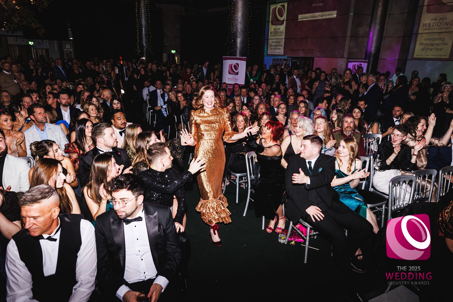 Woman in gold dress walks through applauding crowd at The Wedding Industry Awards gala event.