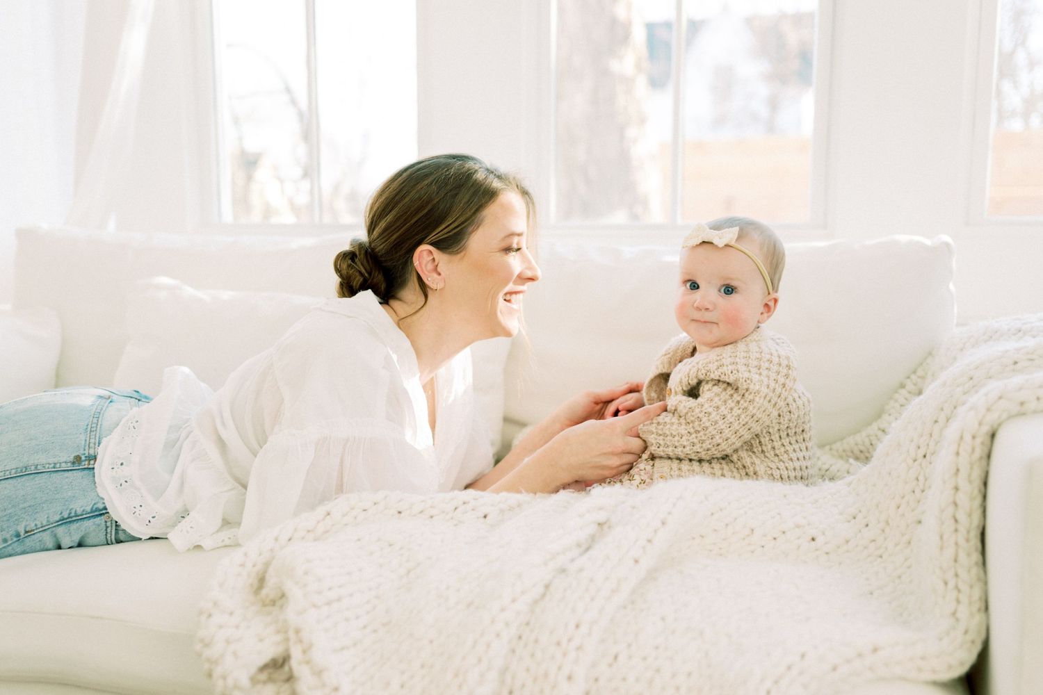 A joyful interaction between adult and baby on a white couch filled with soft cushions and natural light.