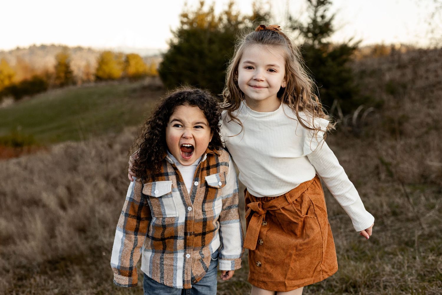 Two young children playing and laughing outdoors on a hillside during autumn with evergreen trees in background.
