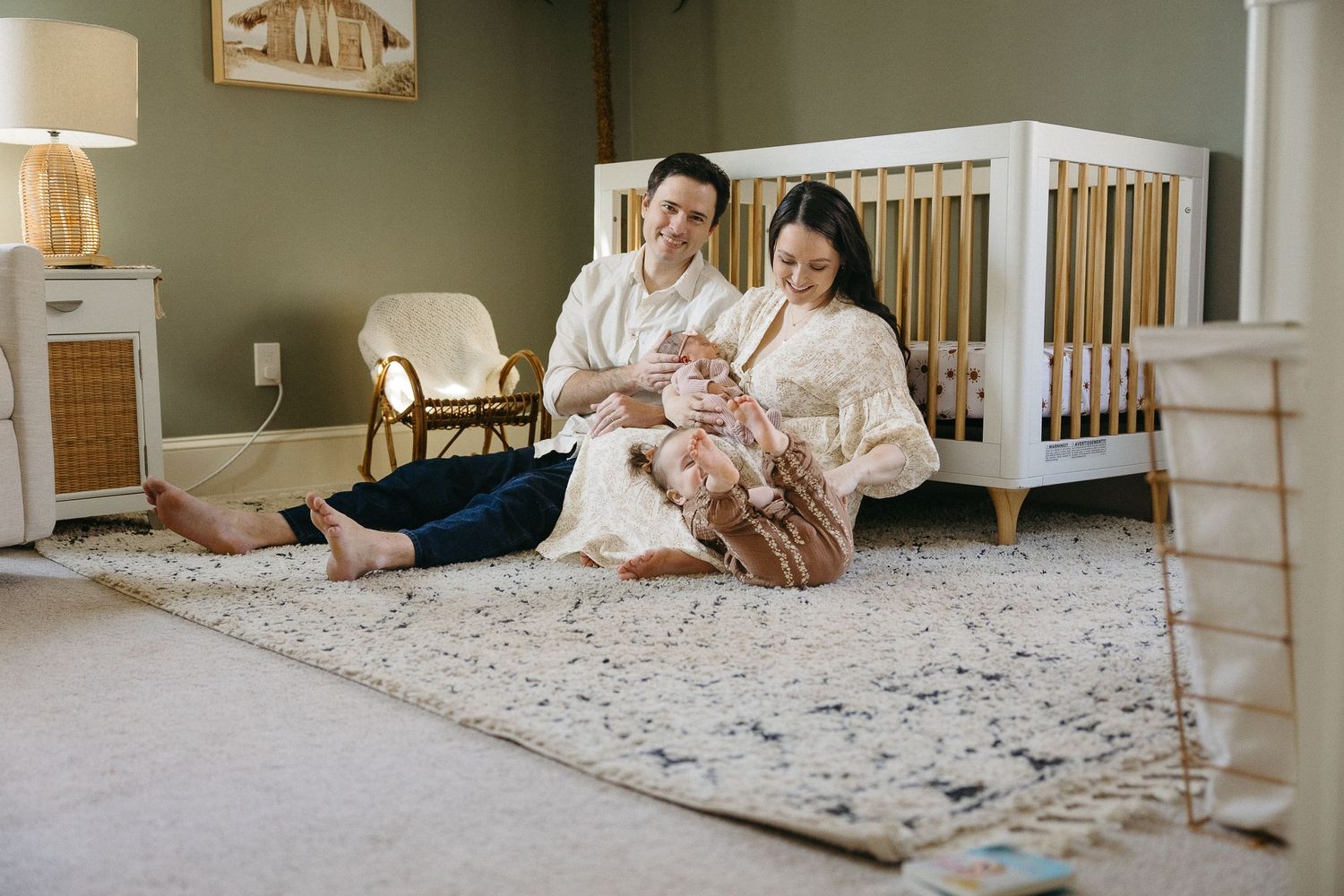 A family sits together on a plush carpet in a cozy nursery with a wooden crib and warm lighting.
