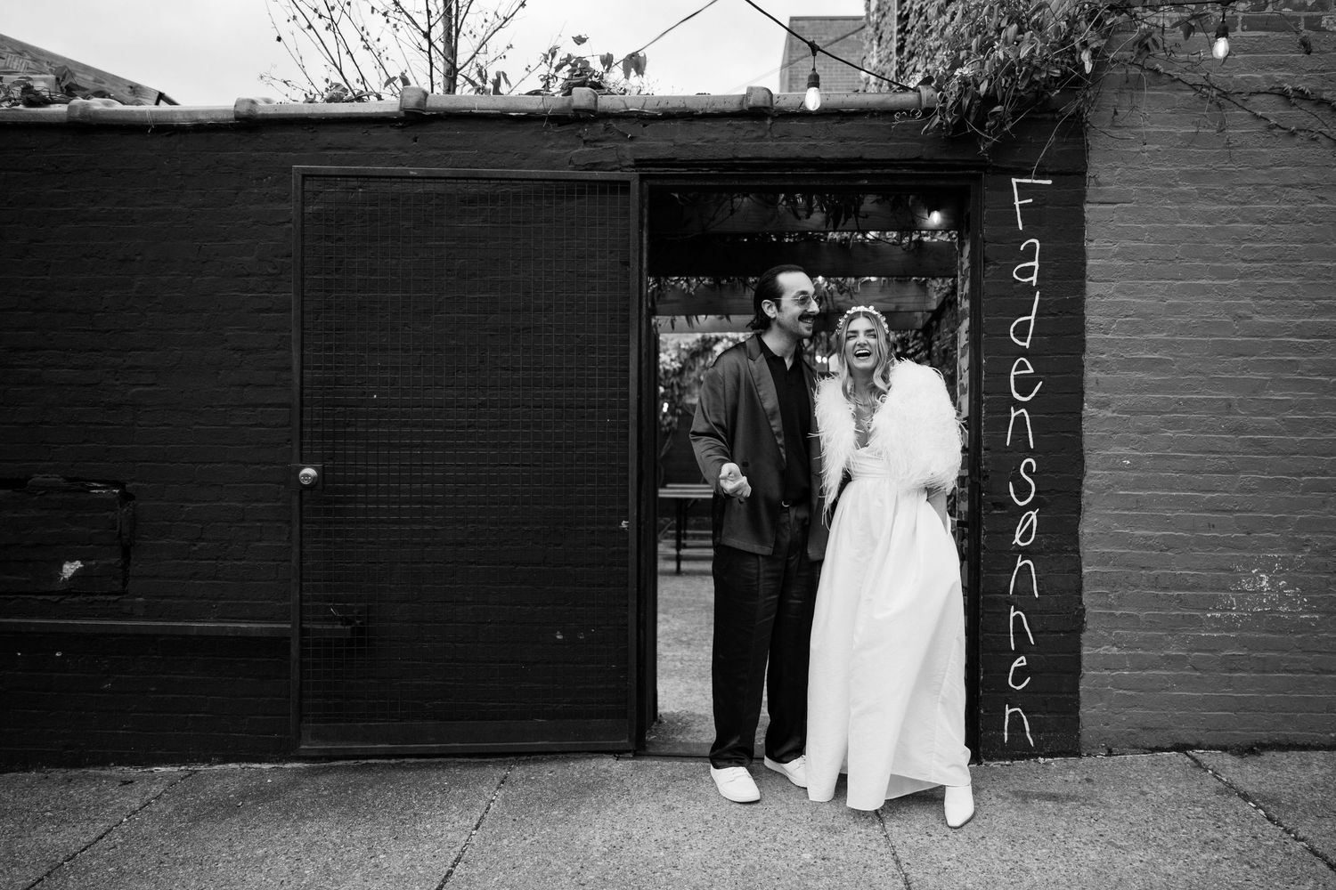 A couple in wedding attire poses in front of a black brick wall with a doorway.