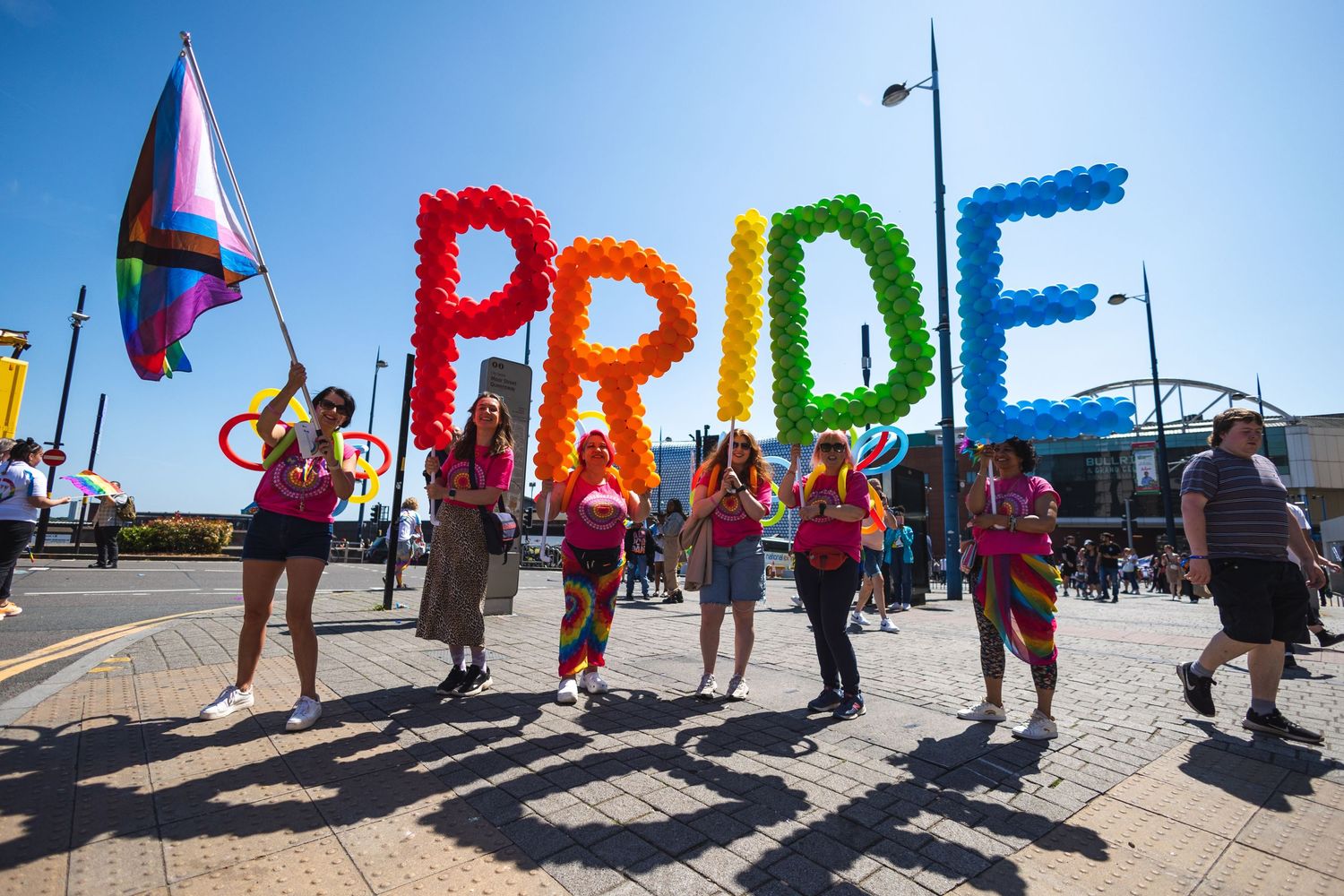 Colorful balloon letters spelling PRIDE carried by parade participants during sunny celebration.