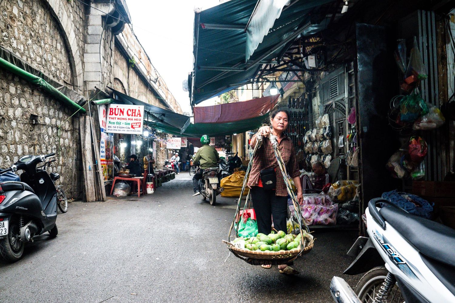 A vendor carries fresh produce in traditional baskets through a narrow street market in Vietnam with parked scooters nearby.