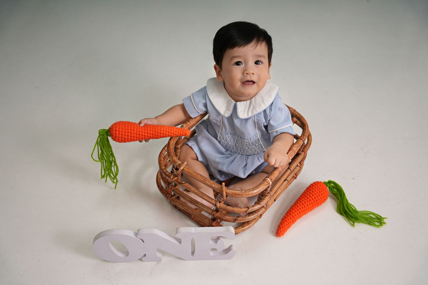 Baby in blue outfit sits in woven basket with decorative orange carrots and ONE text display on white background.