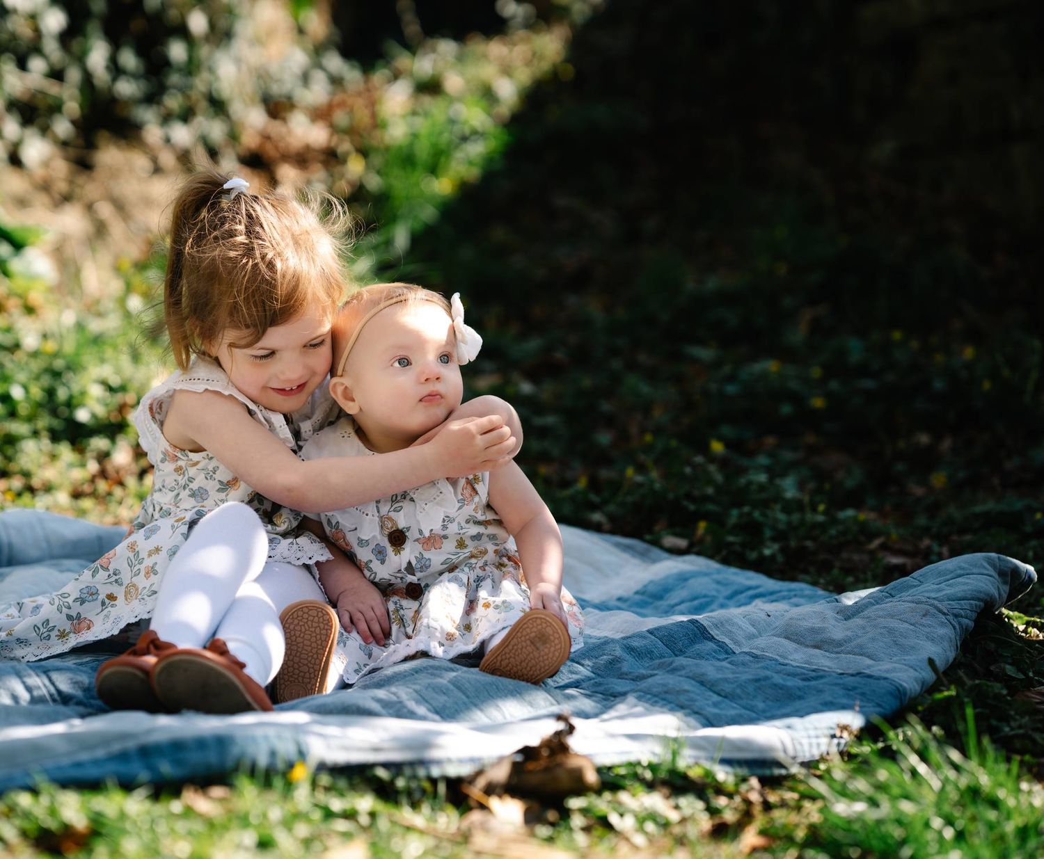 Two small children share a tender moment while sitting on a picnic blanket in dappled sunlight.