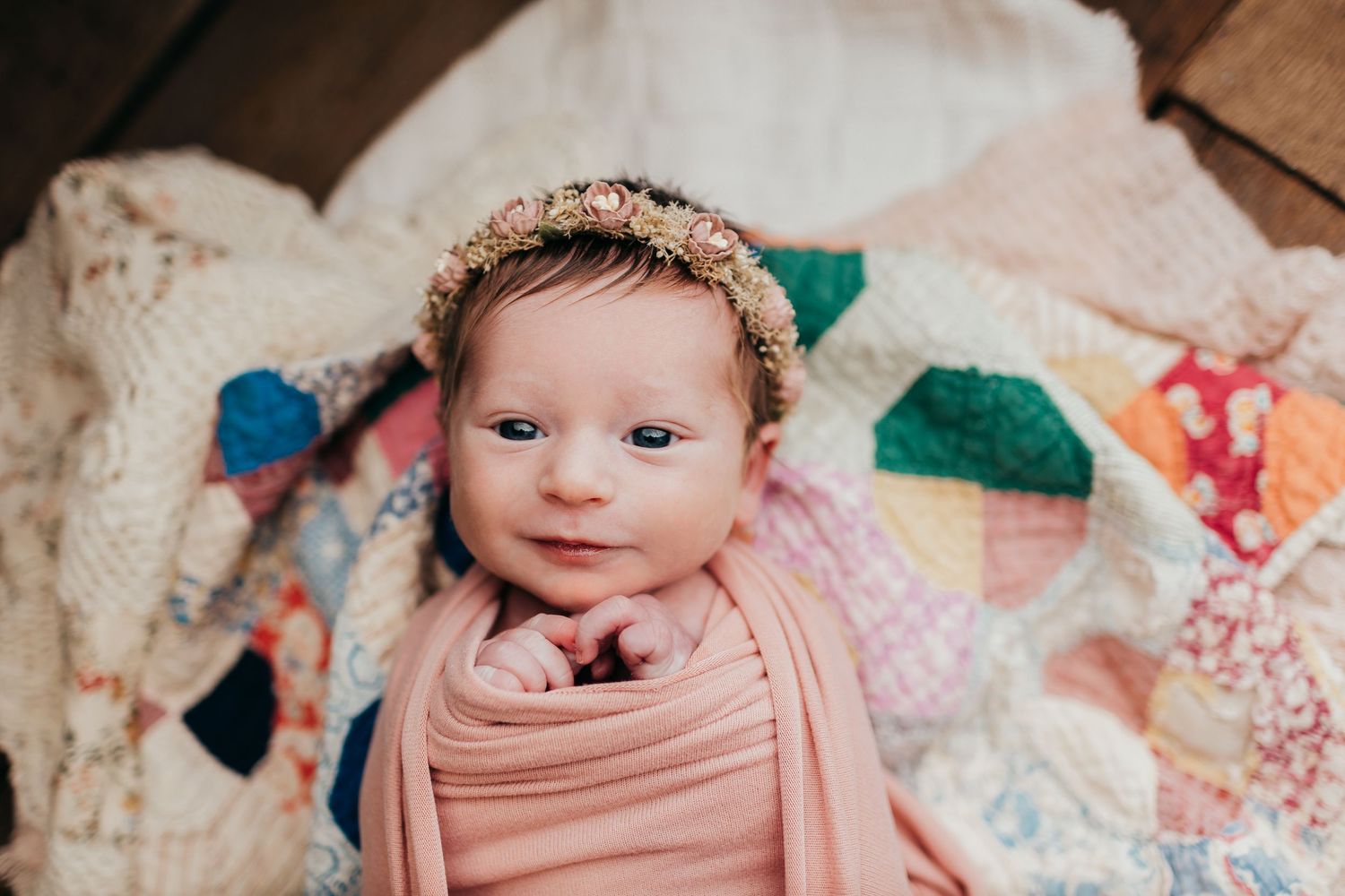 Baby wearing floral crown and pink wrap lying on patchwork quilt during newborn photoshoot.