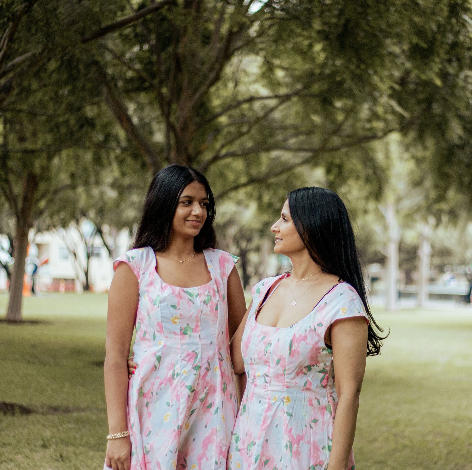 Two women in matching floral pink dresses smile at each other while standing in a sunny park setting.