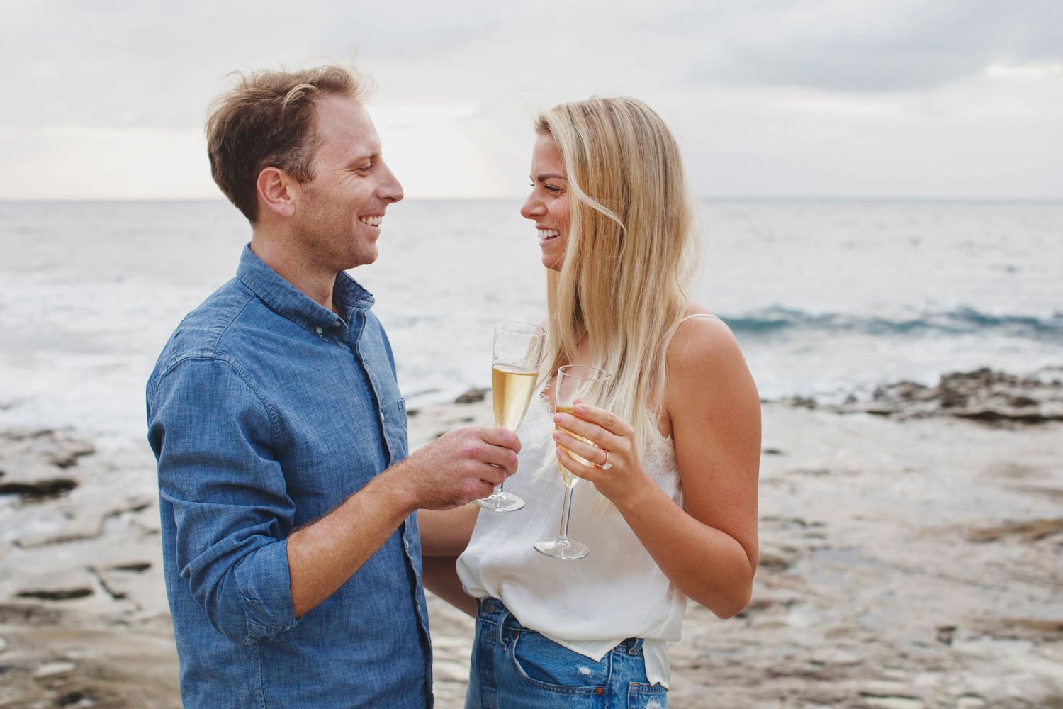 Couple toasting with champagne glasses on rocky beach at sunset.