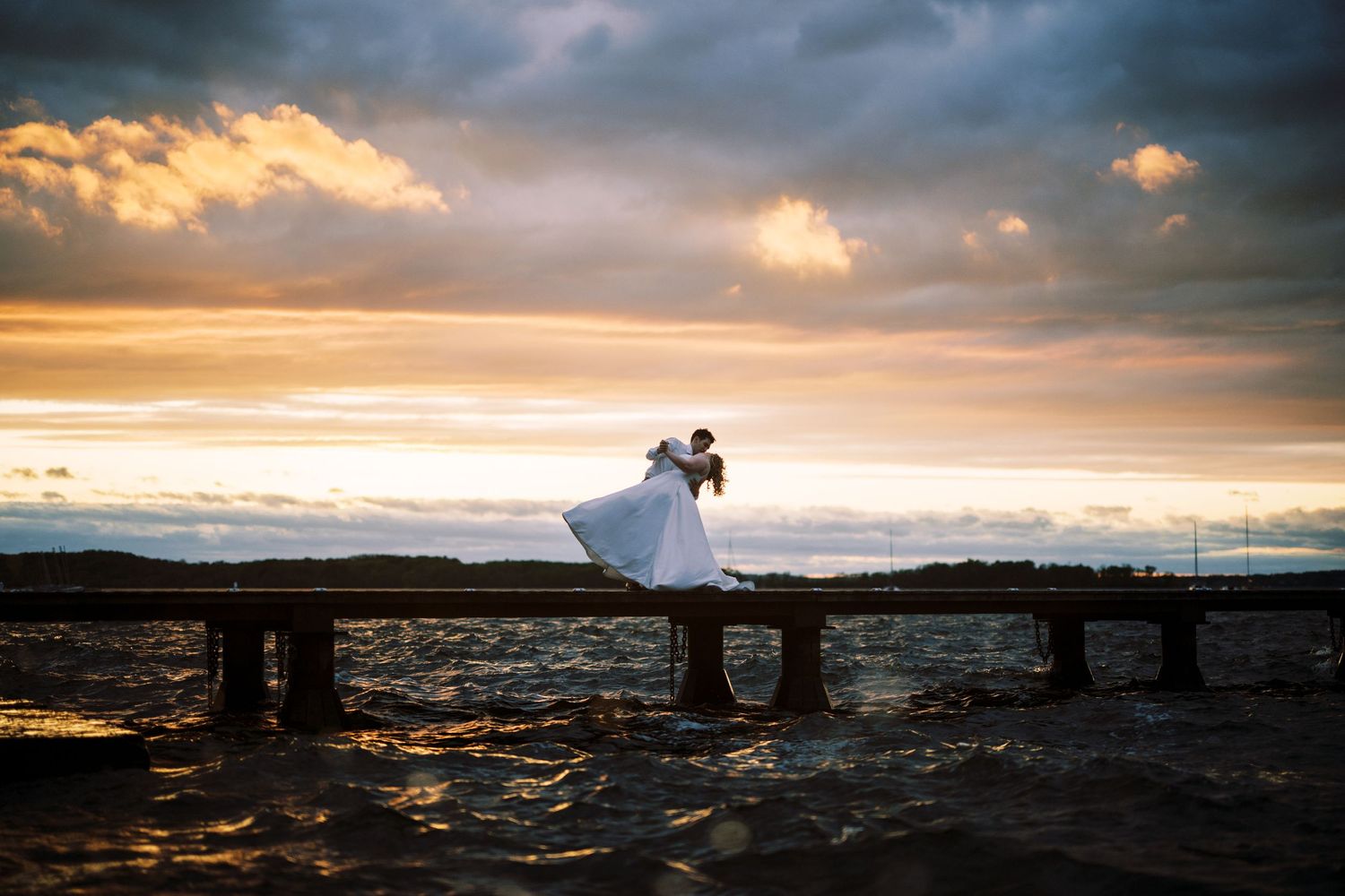 A couple in wedding attire silhouetted on a wooden pier at sunset with dramatic clouds and ocean waves in the background.