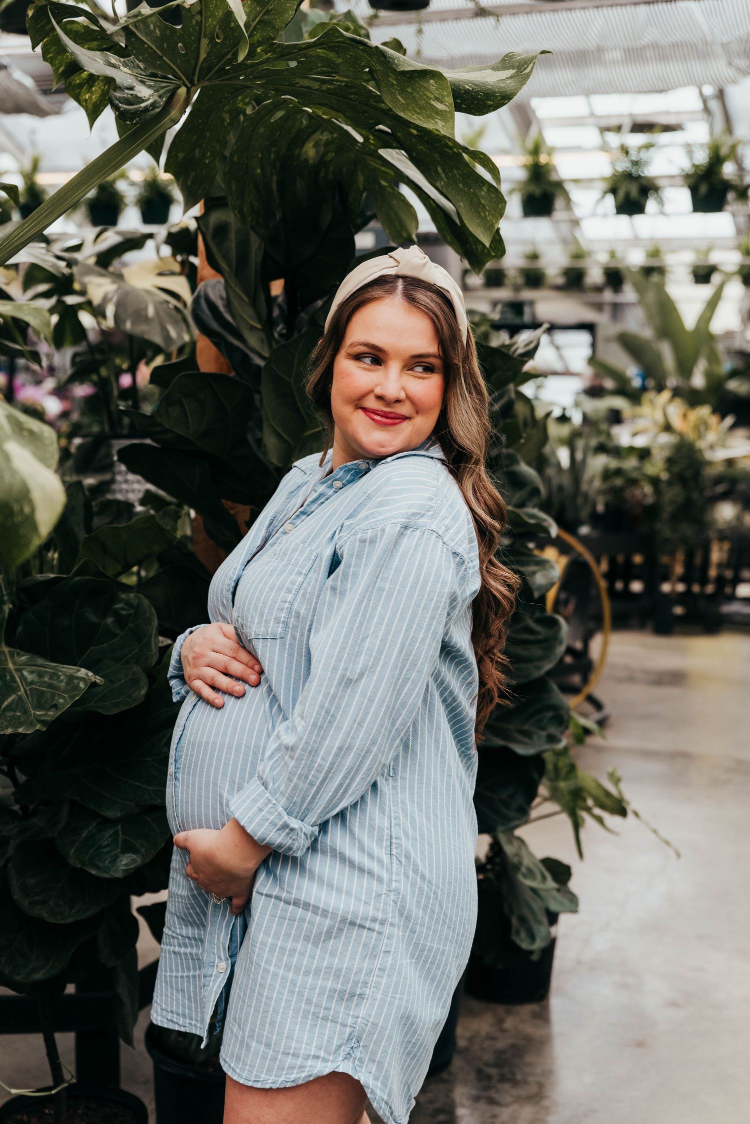 A pregnant woman in a blue striped dress poses in a lush greenhouse at Longfellow's Greenhouse, Manchester, Maine for her maternity session.