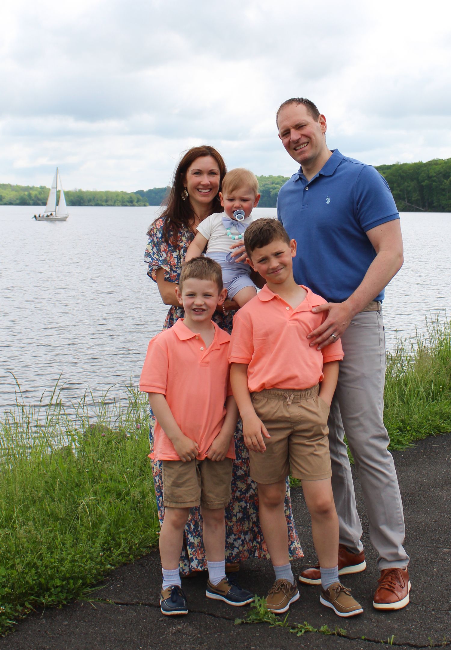 Family standing together by lake with sailboat in background.