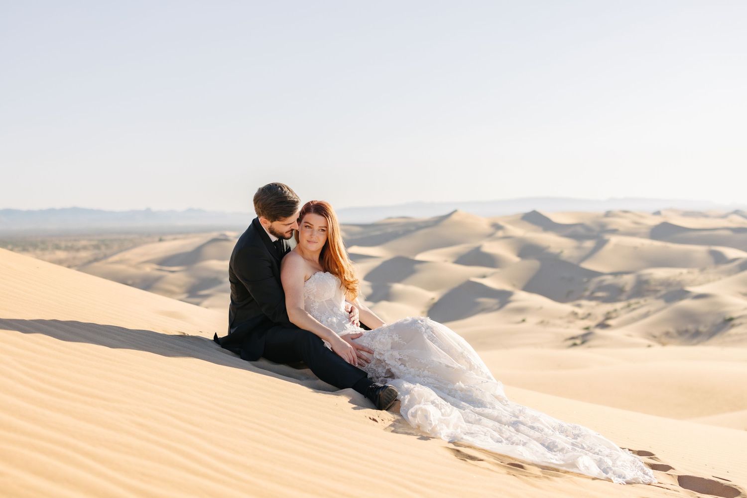 Couple in wedding attire sitting together on a sand dune overlooking desert landscape.