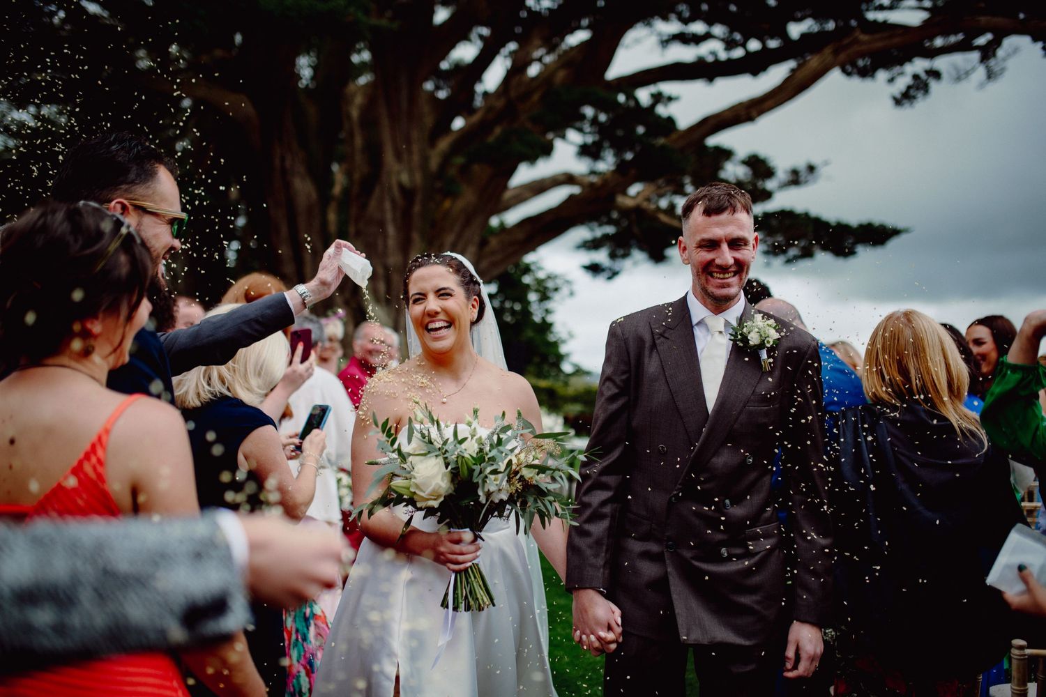 Joyful wedding exit with guests showering the couple with confetti under a large tree.