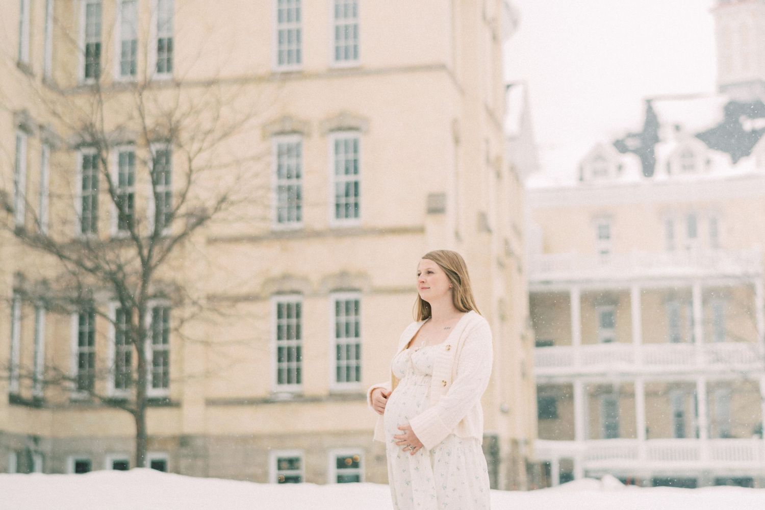 Expectant mother in white dress stands in winter scene by historic stone building during light snowfall.