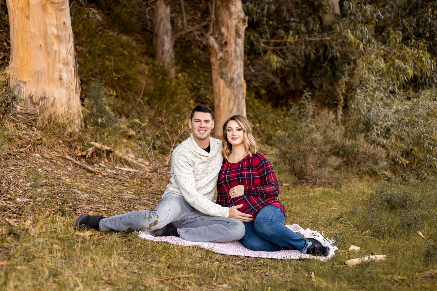 A couple sits on a blanket in a forest, the woman cradling her baby bump while the man holds her belly, both smiling warmly.