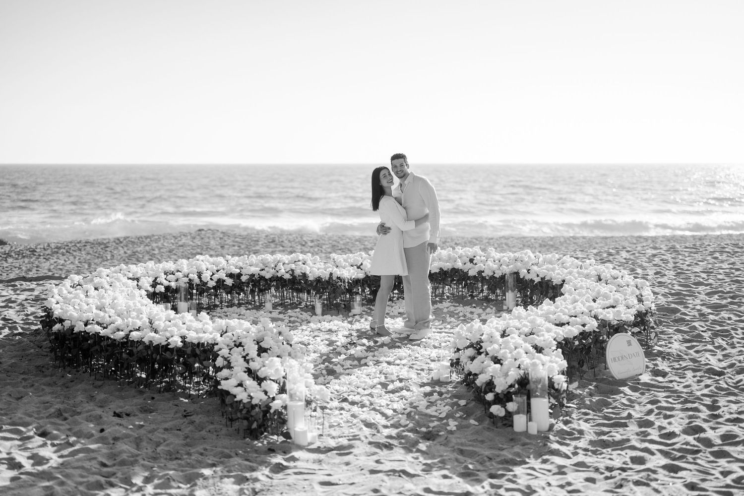 A romantic seaside setup with a heart-shaped floral arrangement on the beach at sunset.