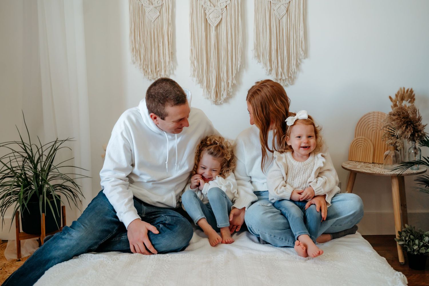 A family sits together on a white bed, wearing casual denim and white clothing, with macrame wall decor behind them.