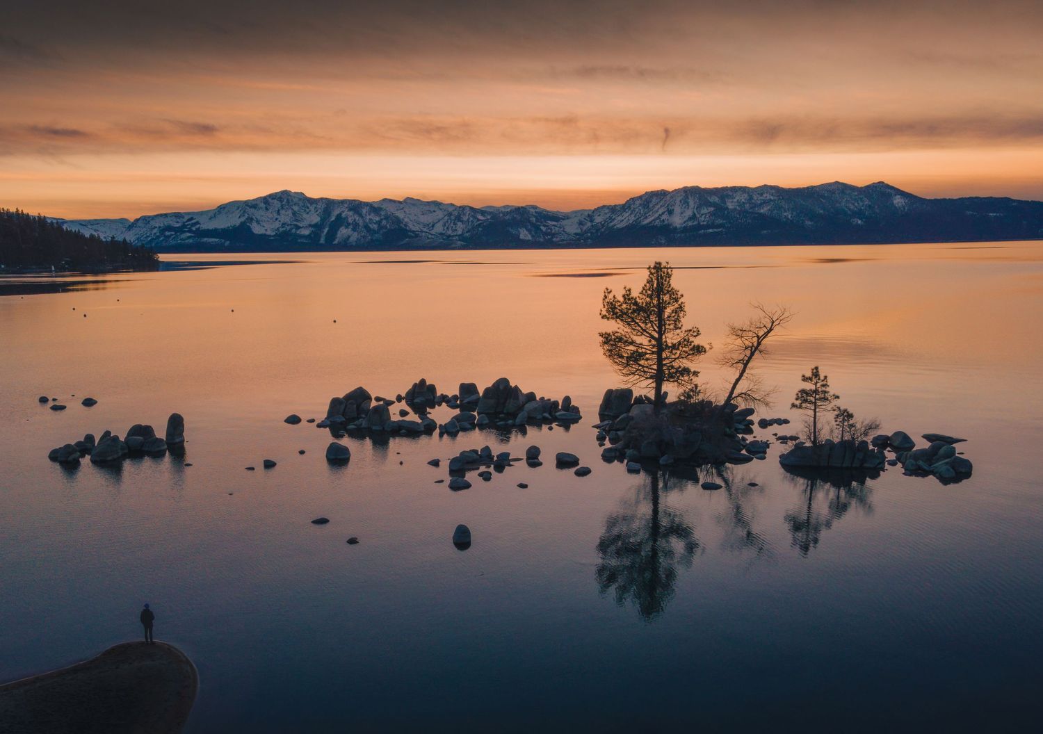 Silhouette of trees on rocky outcrop in Lake Tahoe during golden sunset with snow-capped mountains in background.