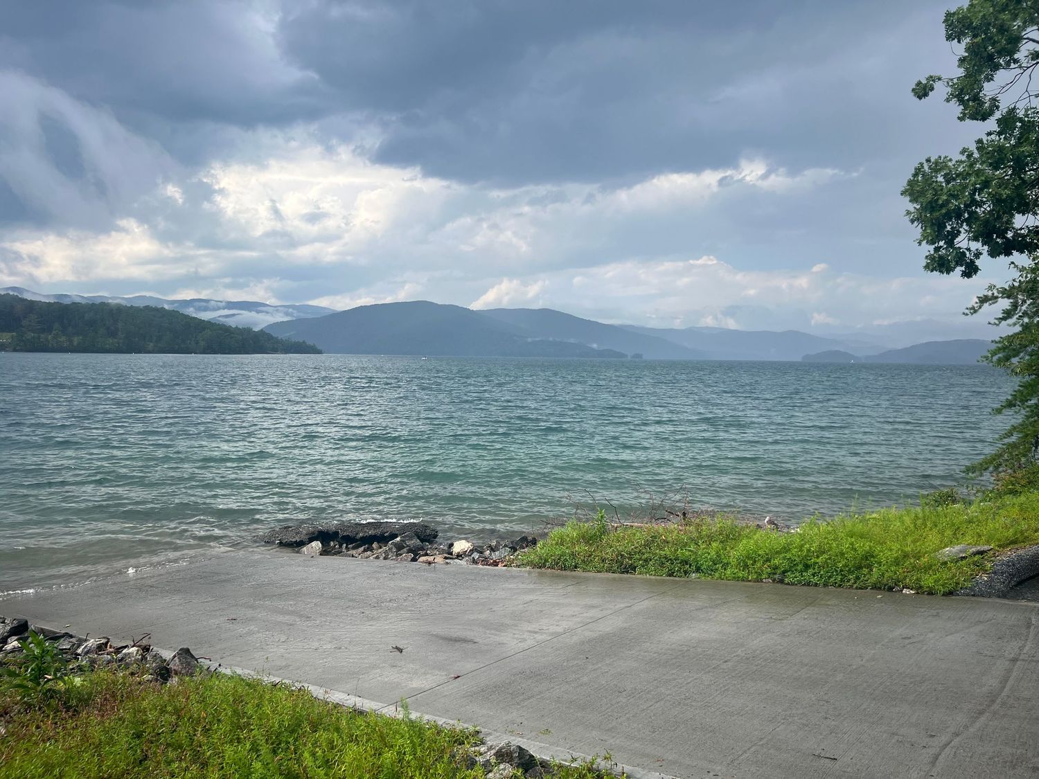 Choppy turquoise lake with rocky shoreline, concrete boat ramp, green hills, and dramatic stormy sky with mountains in background.