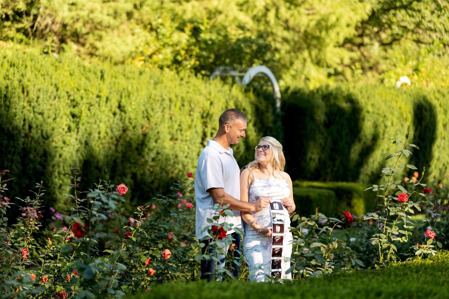 Couple smiling at each other in a lush garden surrounded by red roses and green hedges on a sunny day.