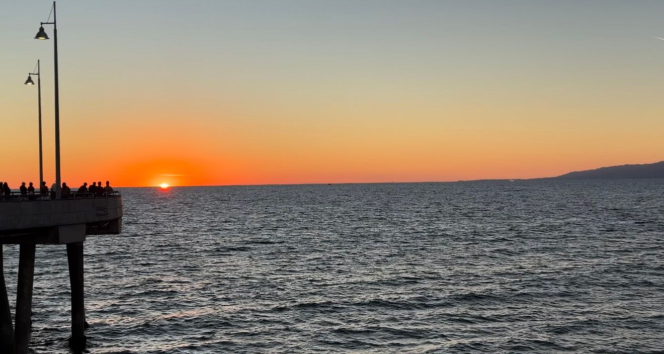 Orange sunset glowing over calm ocean waters with silhouetted people and lamp posts on a pier in the foreground.
