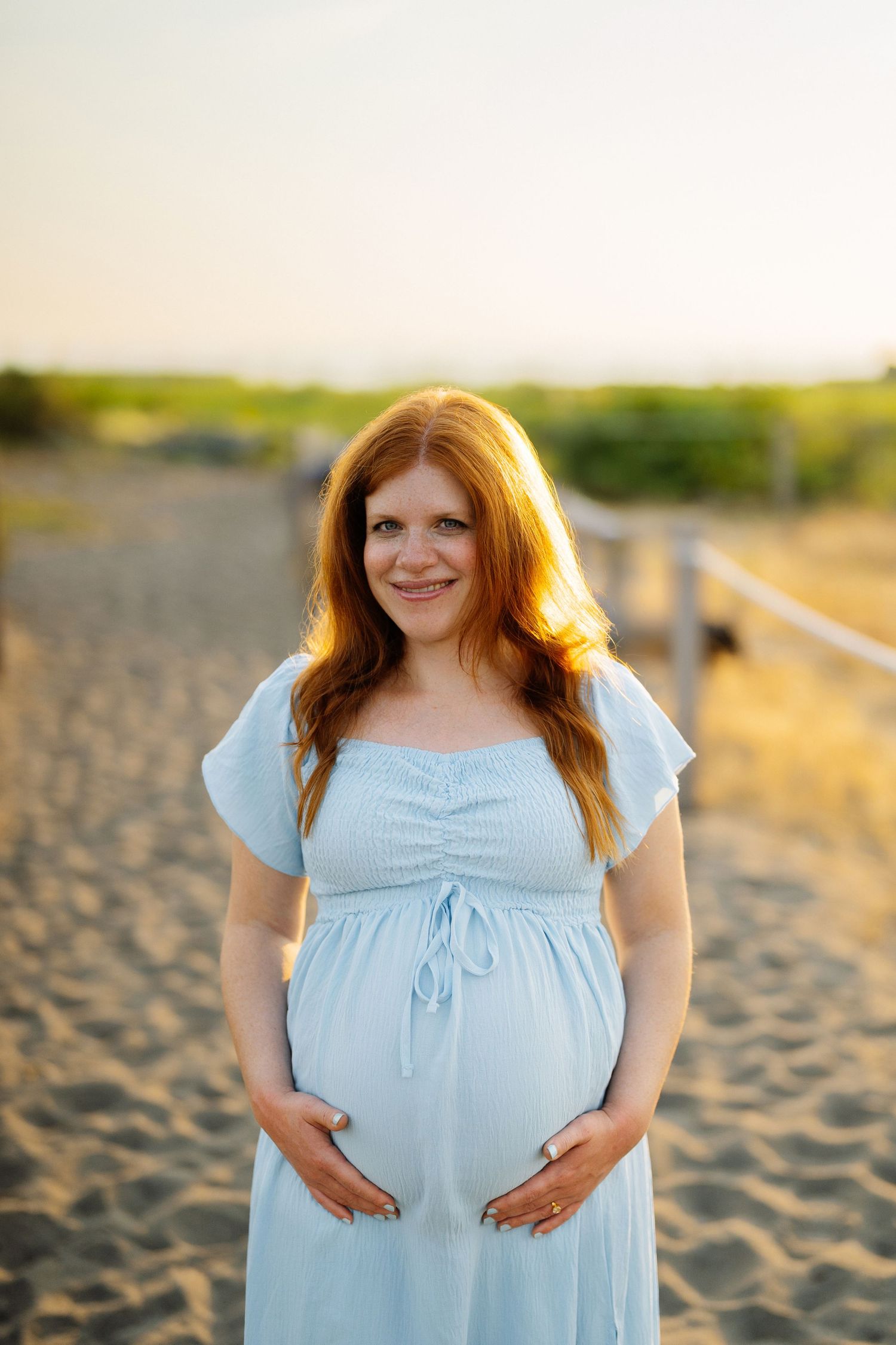 Smiling pregnant woman with long red hair in a light blue dress cradles her baby bump on a sandy beach at golden hour.
