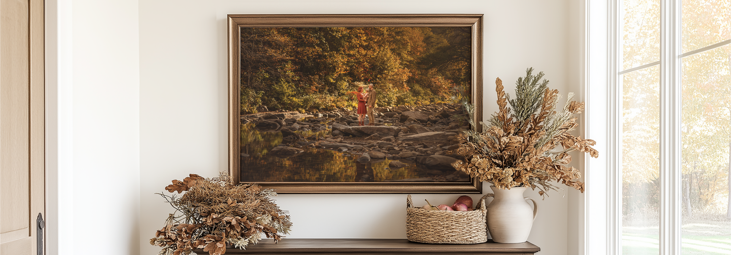 Framed autumn landscape painting above a wooden console table with dried floral arrangements in warm fall tones.