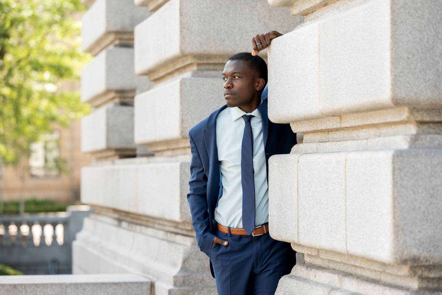 A young Black businessman in a navy suit and tie leans against a stone building column outdoors.