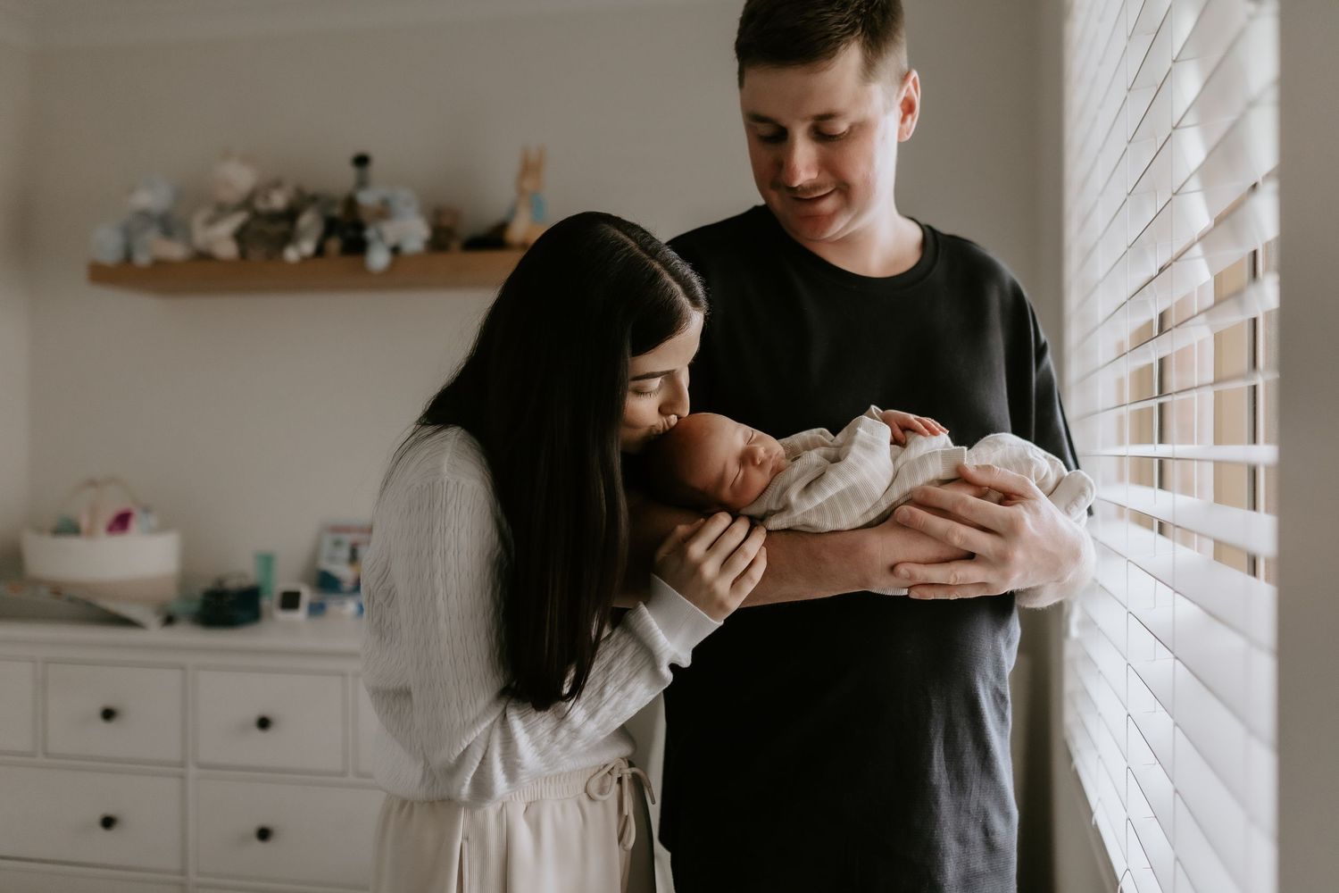 Young parents lovingly holding their newborn baby in a bright nursery room near a window.