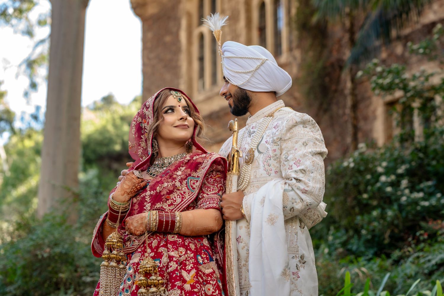 Indian bride in red lehenga and groom in white sherwani pose together at an outdoor wedding venue.