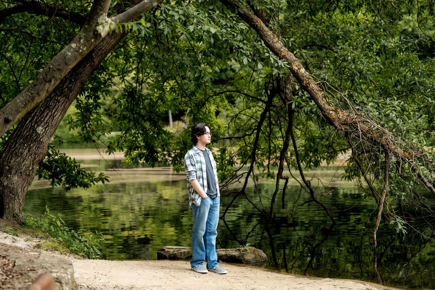 A young man stands by a tranquil green pond beneath arching tree branches on a sandy path.