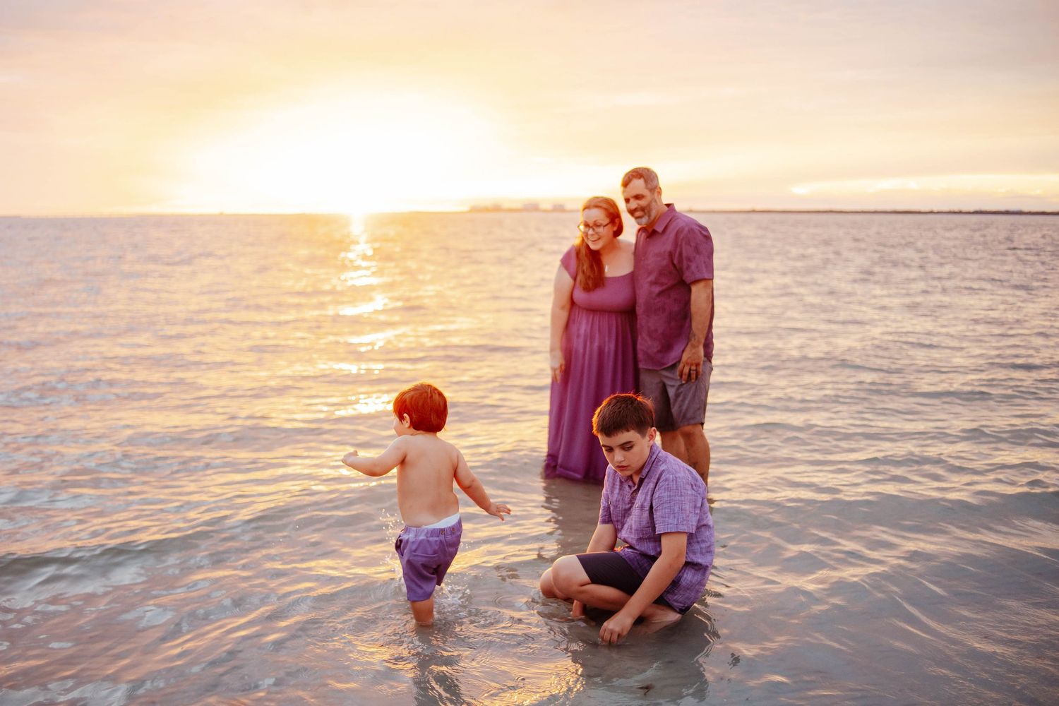 Family of four in coordinating purple outfits poses in shallow ocean water at sunset, with two young children playing near the shore.