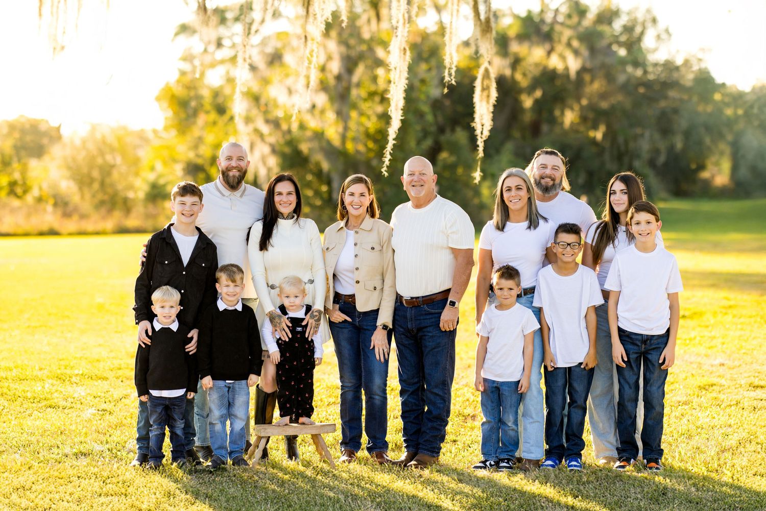 Large multigenerational family posing together outdoors on green grass with trees and golden sunset light behind them.