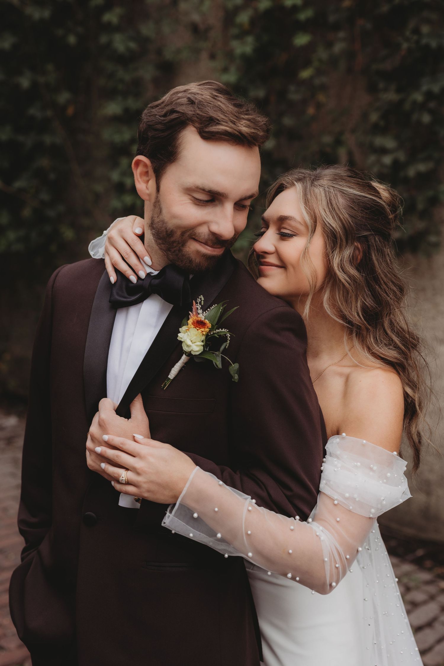 A bride in a white off-shoulder gown embraces a groom in a burgundy tuxedo outdoors.