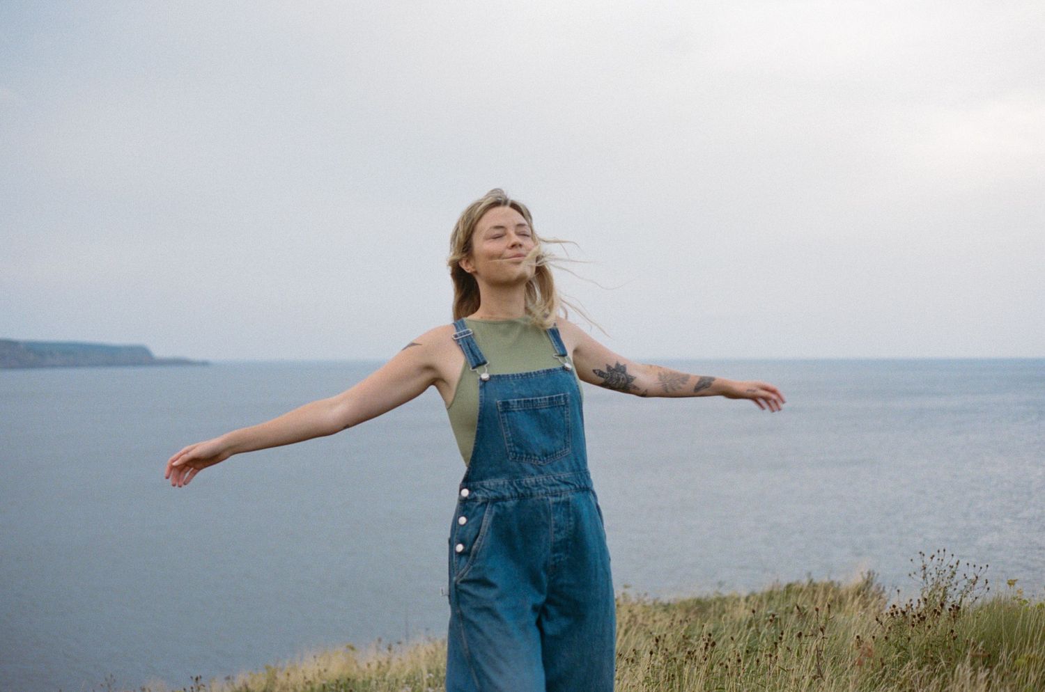 blonde woman with tattoos spins around in a meadow by the ocean during Self Love Session