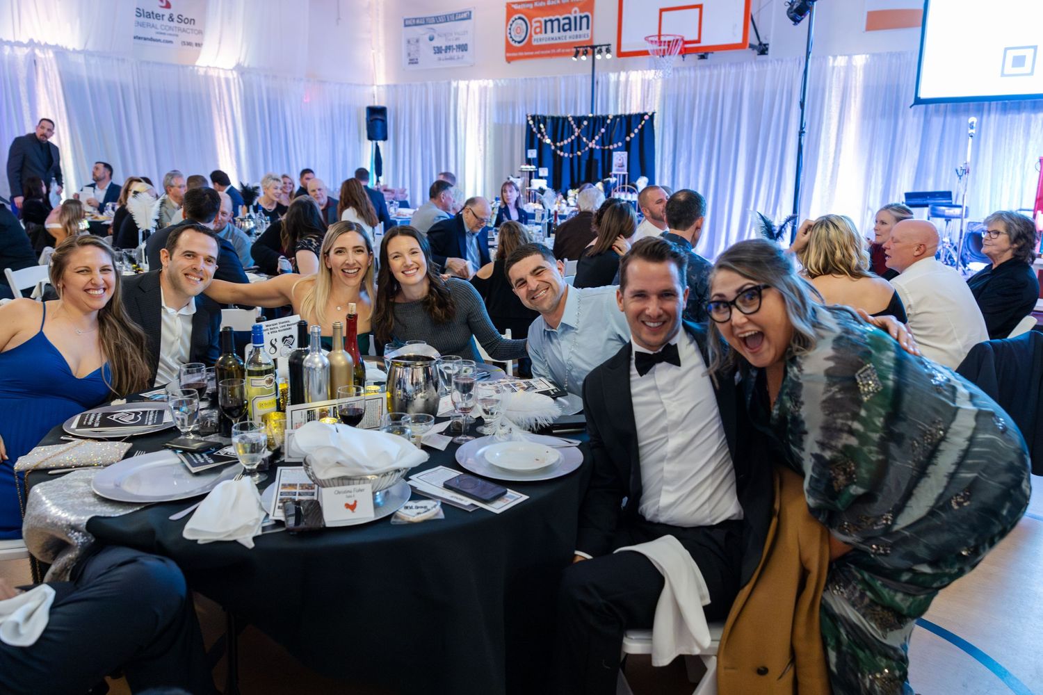 Group of guests celebrating at round dinner tables in decorated gymnasium venue.